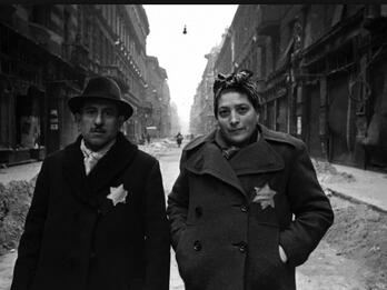 Photograph of man and woman standing in the middle of a city street wearing jackets with yellow Stars of David on breast.