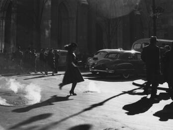 Midtown Manhattan Photograph of woman in a dress and hat walking across a city street with cars, people, and buildings in the background.