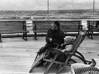 Photograph of man in a skullcap kneeling next to a child sitting in a wooden chair on boat deck, gazing into the distance to the left of the viewer.