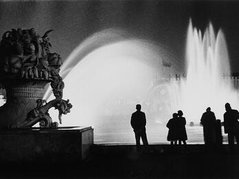 Stone Flower Fountain, Moscow Photograph of a cornucopia statue in the left foreground silhouetted against a large fountain in the background. There are several people standing at the edge of the fountain looking away from the camera toward the water.