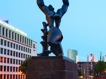 Photograph of bronze memorial sculpture on a rectangular pedestal depicting stylized human figure with hole in chest and abdomen, leaning against a tree stump with its hands in the air and head thrown back, and city buildings in background.