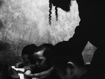 Photograph of man in a hat and sidelocks bending over a desk where three boys are reading Hebrew. 