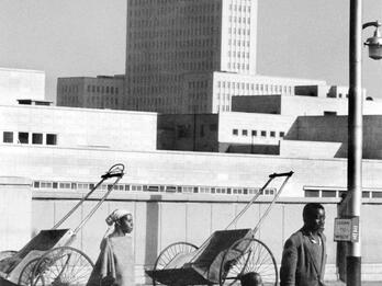 Arriving Family, King George Street, Johannesburg Photograph of man, woman, and child walking down city street with high-rise building and two rickshaws in background.