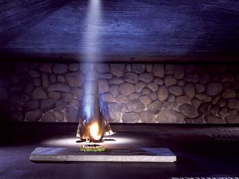 Hall of Remembrance at Yad Vashem Memorial depicting flame burning from a base and illuminating a wall with an angular roof and basalt walls, with place names in English and Hebrew on the floor.