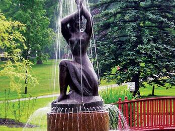 Spirit of the Sea, Library Square Park, City of Bath, Maine Bronze sculpture and fountain in the shape of a partially nude woman, resting on one knee and raising her arms above her head, set in a park with trees and a bridge in the background.