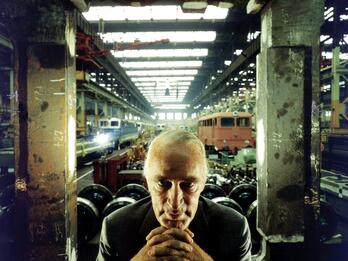 Arnold Krupp Photograph of man wearing a suit seated in warehouse facing camera with chin resting on folded hands, framed by two concrete pillars.