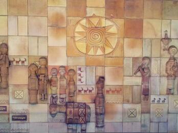 Ceramic relief made up of squares featuring stylized figures, animals, geometric shapes, and sun in center above figures.