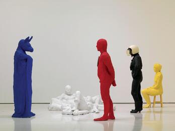 The Costume Party Mixed media sculpture featuring six figures in various poses: a male and a female figure reclining on the ground, a woman seated in a chair to the right, a man with a donkey mask standing to the left, Superman standing in center, and a woman standing in the background.