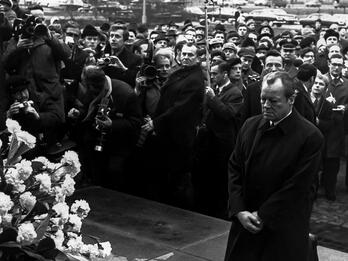 West German Chancellor Willy Brandt Kneels in Front of the Jewish Heroes Monument Paying Tribute to Jews Killed by the Nazis during the 1943 Uprising in the Warsaw Ghetto, December 7, 1970 Photograph of a man in a long coat kneeling in front of a memorial wreath on a pedestal as crowd watches and photographers take photos.