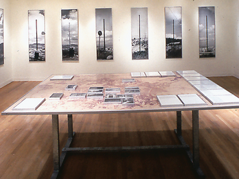 The Eruv Installation showing set of eight photographs arranged on back wall, and table in foreground with fourteen photographs and fourteen texts lying flat.