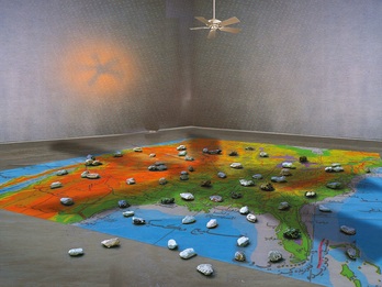 Art installation of large map on floor with rocks in various places and ceiling fan overhead. 