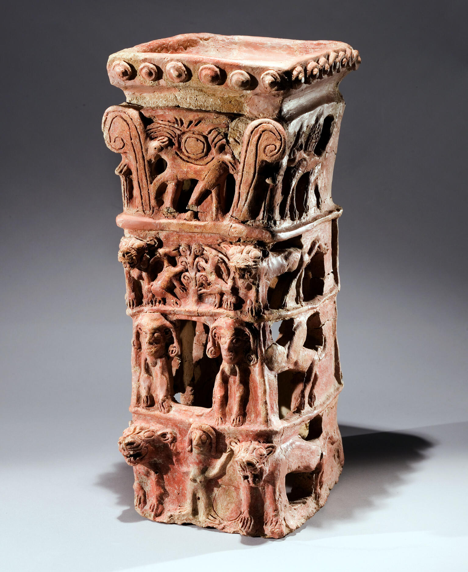 Ceramic offering stand with flat top and four layers of decoration: bottom layer of nude female with raised arms flanked by lions, second layer of two winged figures, third layer of goats eating leaves off tree, and fourth layer of four-legged animal with winged sun disk and palm fronds.