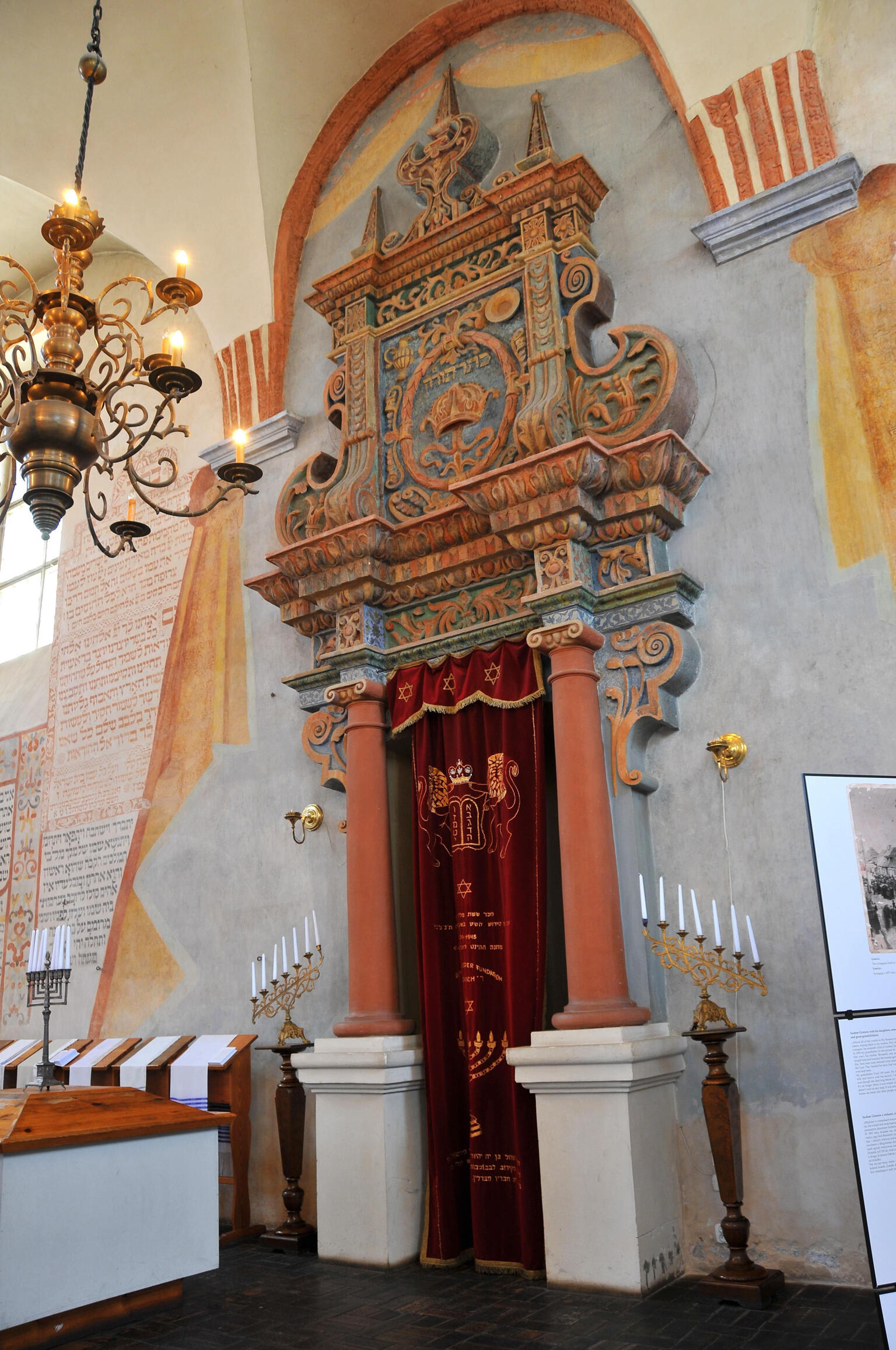 Photograph of carved doorway set in wall with curtain across it, surrounded by candelabra, Hebrew writing on wall, and chandelier in forefront. 