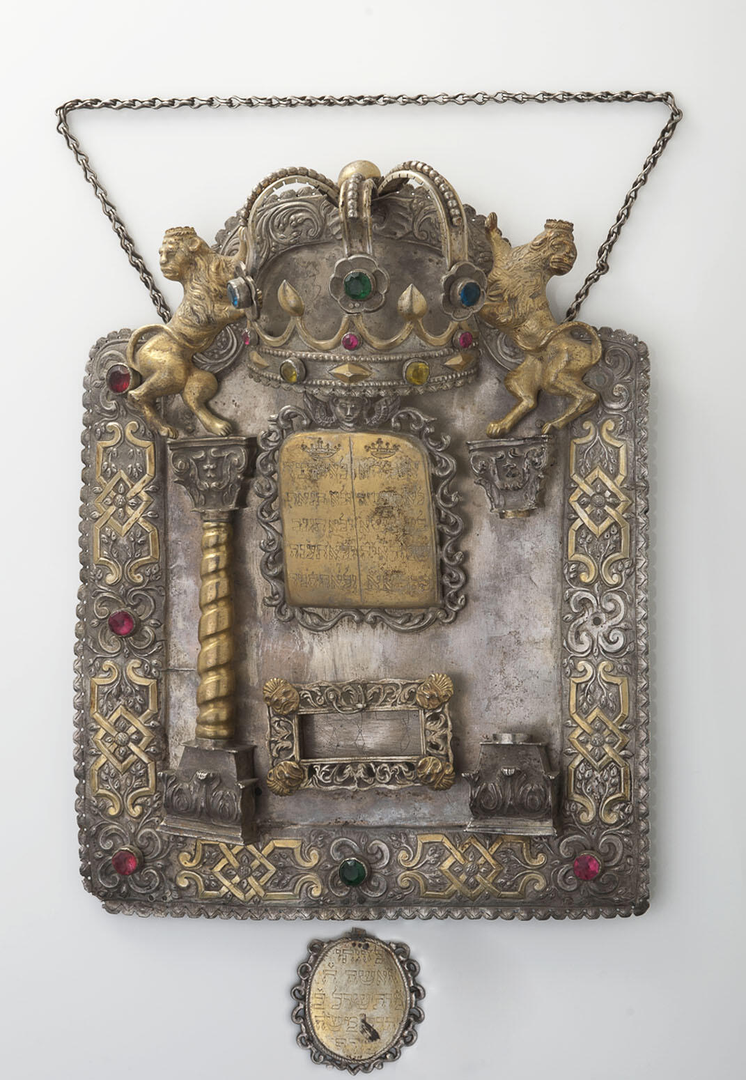 Shield with decorative border inlaid with semi-precious stones and adorned with lions and crown at top, column on left and plaque in middle with Hebrew text. Shield with decorative border inlaid with semi-precious stones and adorned with lions and crown at top, column on left and plaque in middle with Hebrew text.