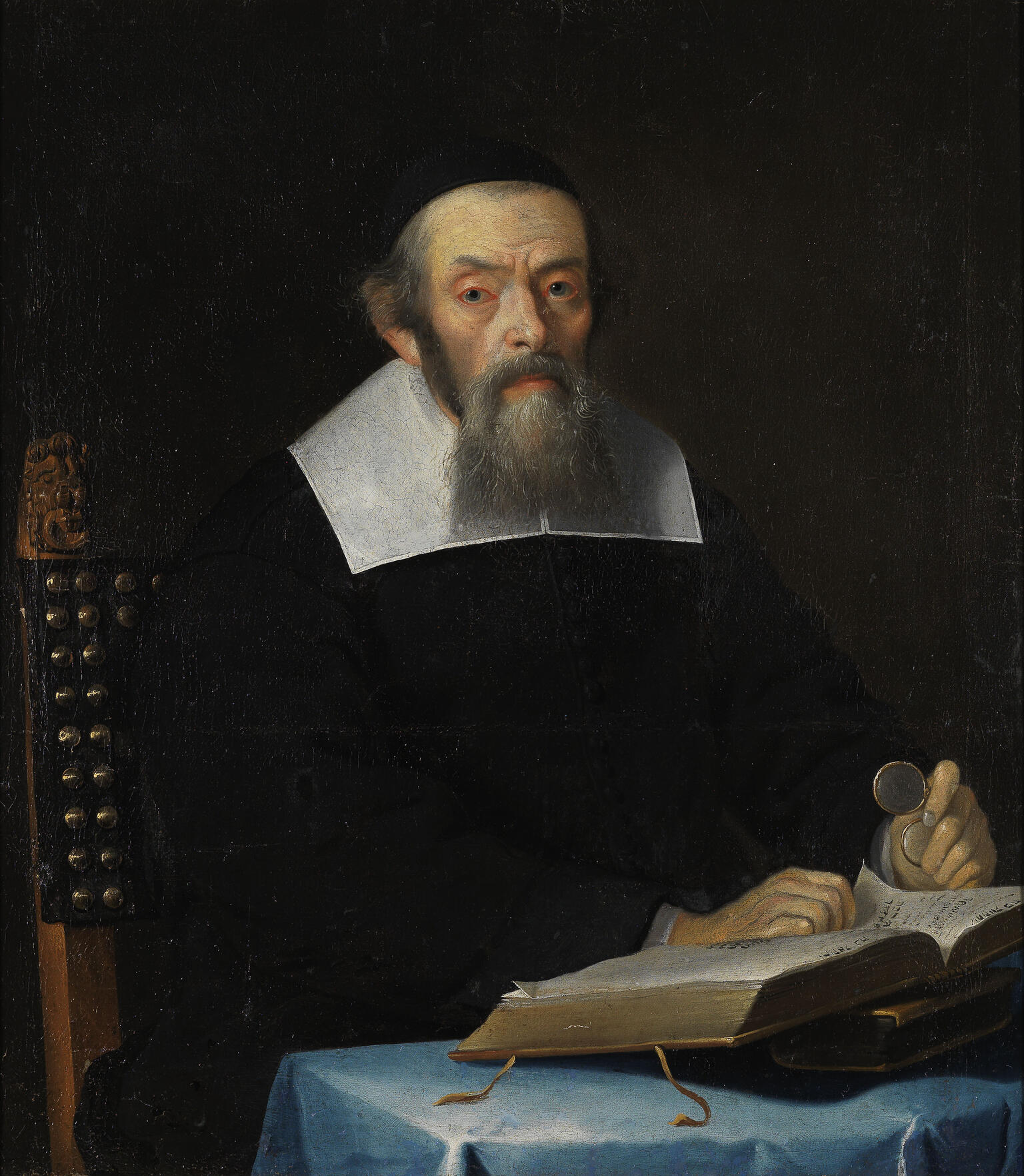 Portrait painting of man in robes with collar facing viewer and sitting at a desk, his hands resting on an open book and holding glasses in his left hand. 