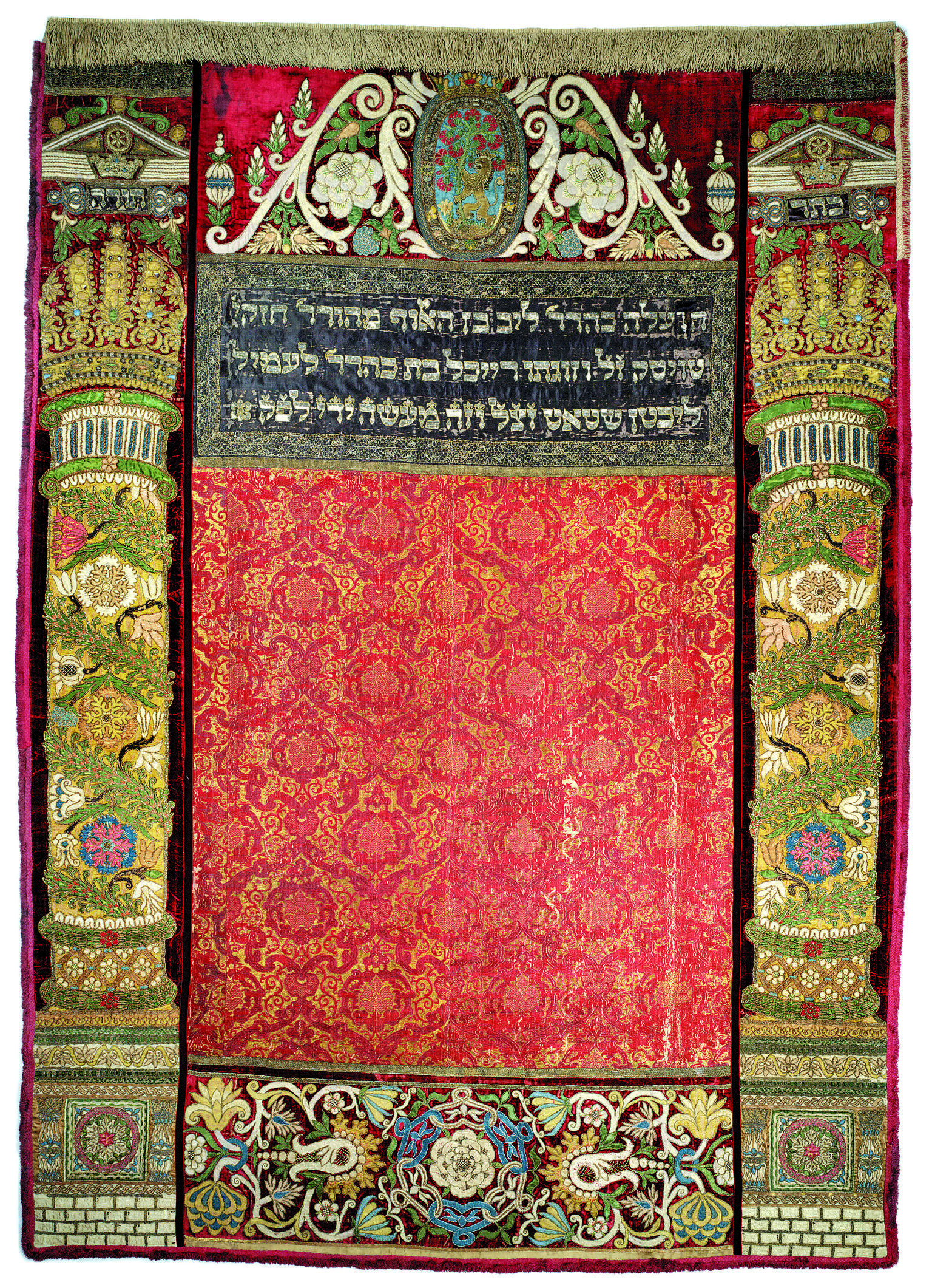 Embroidered curtain of columns with crowns on left and right sides and decorative floral motifs throughout, and Hebrew text in top center.