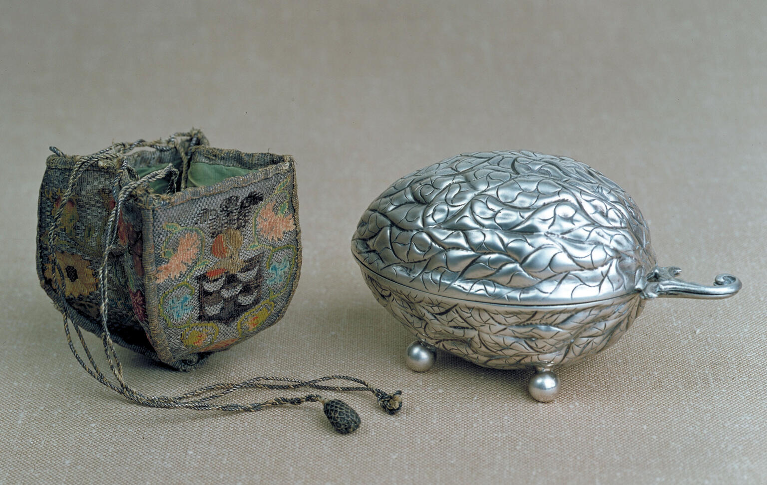 Silver oval container of decorative leaf design and woven, embroidered case next to it.