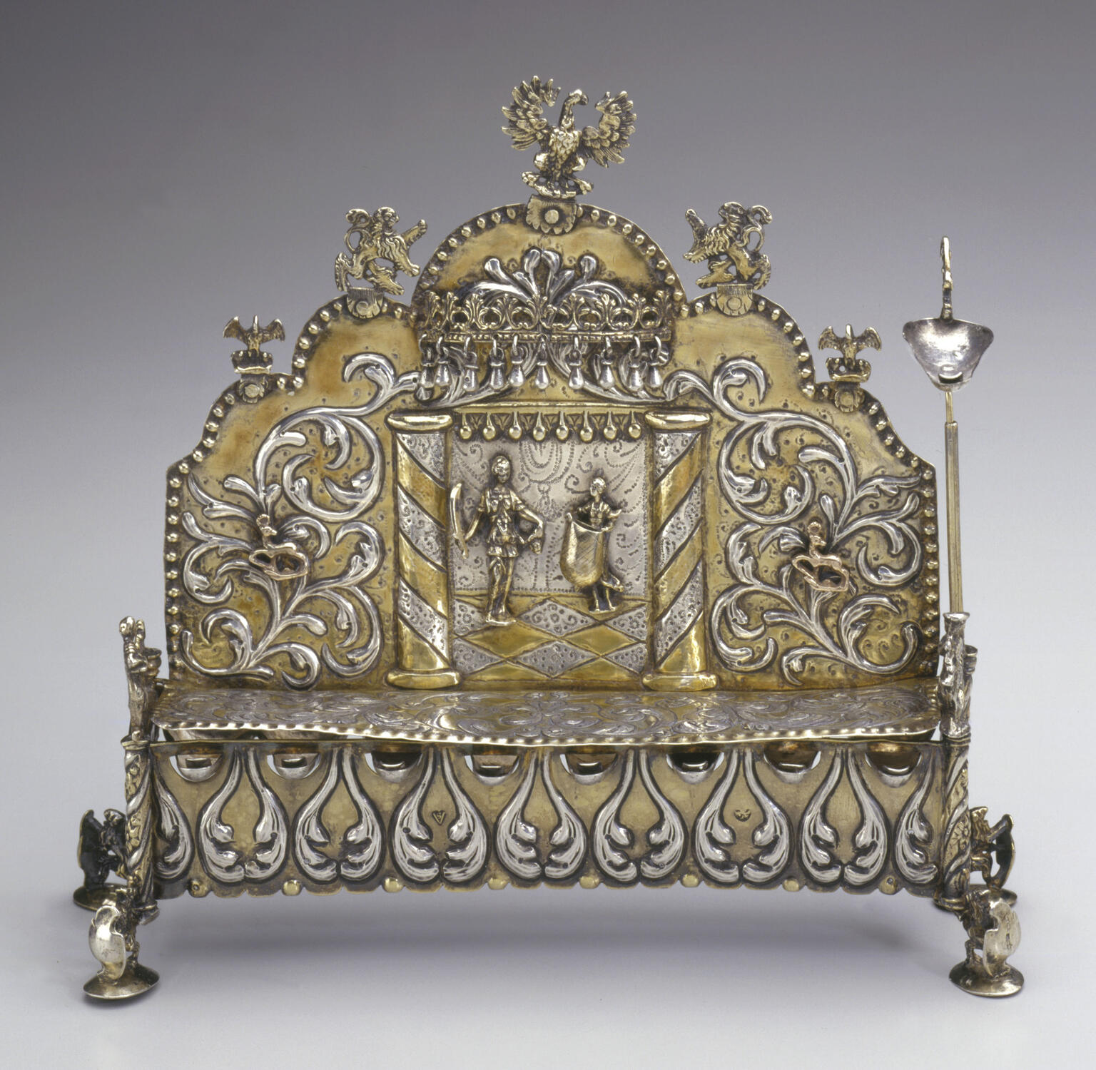 Silver oil lamp with row of eight cups on bottom, and elaborate carvings of two figures and floral designs on lamp with carved birds on top. 