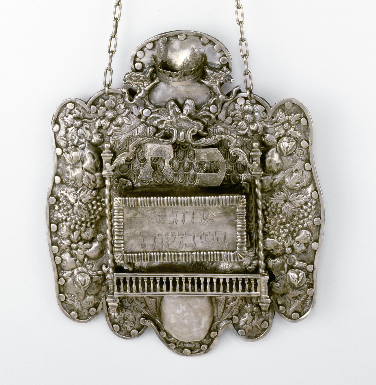 Engraved shield of flowers, grapes, and other decorative elements around rectangular Hebrew inscription. Engraved shield of flowers, grapes, and other decorative elements around rectangular Hebrew inscription.