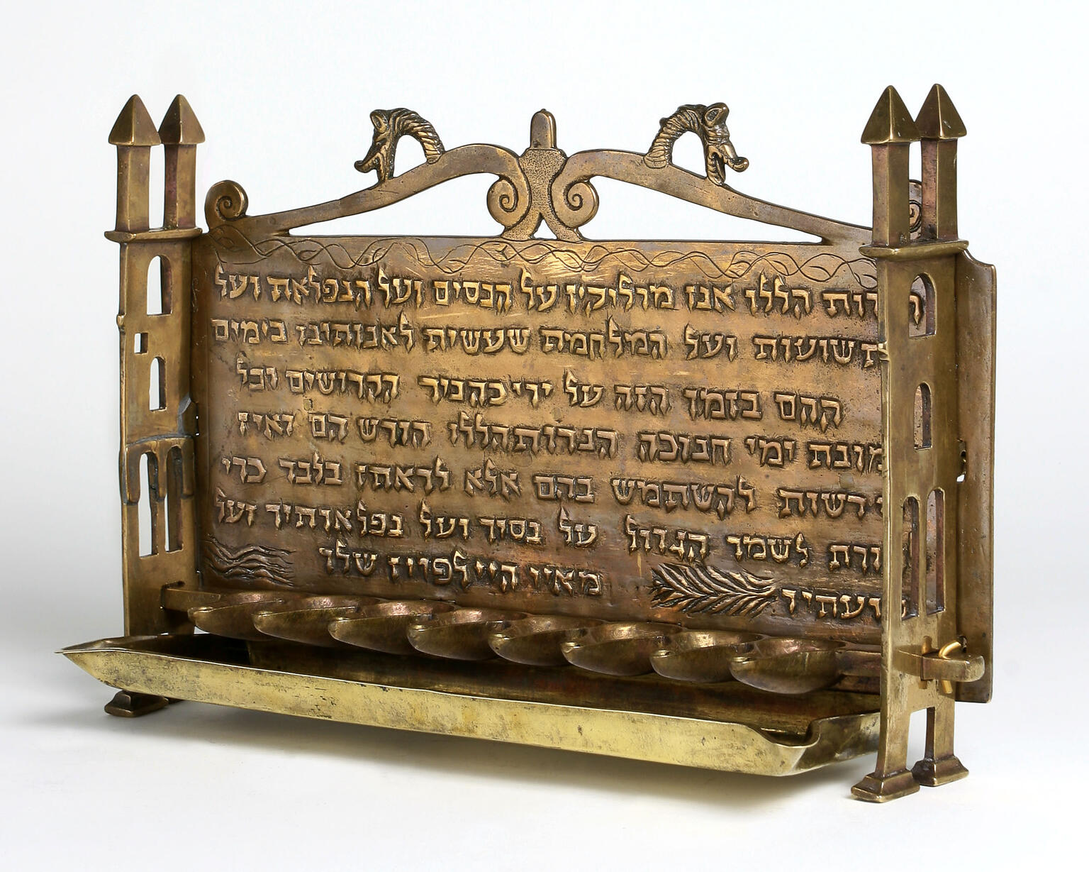Oil lamp with row of eight cups at the bottom carved with Hebrew inscription and animal heads, and sides resembling buildings.
