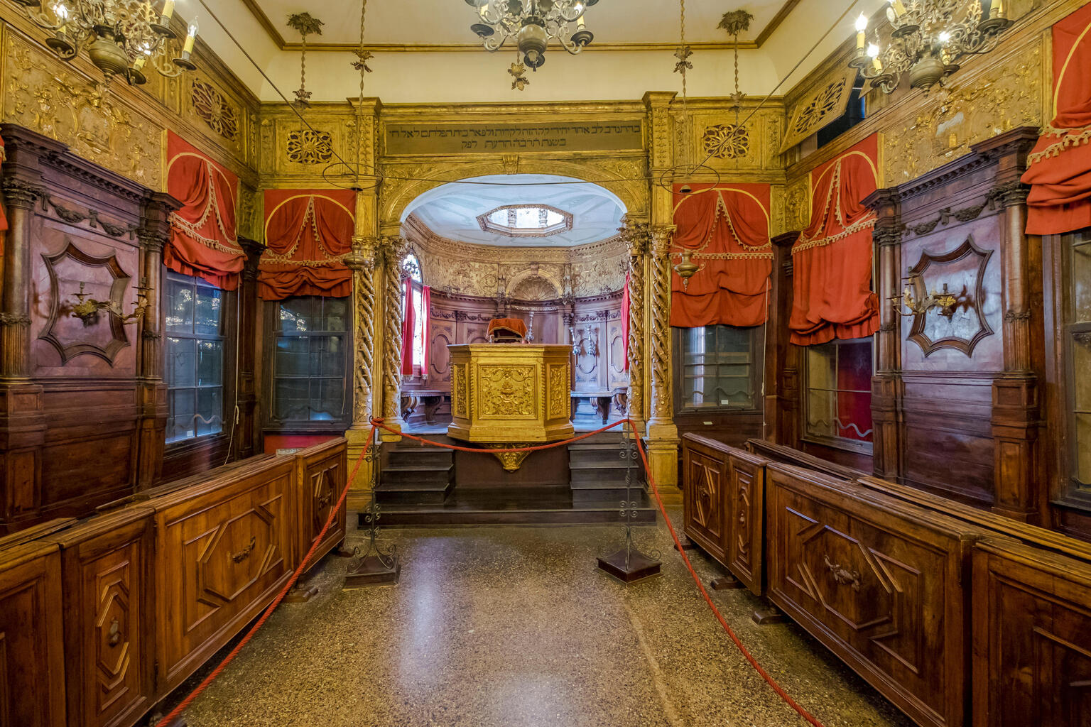 Photograph of room lined with narrow wooden pews and set of stairs at far end of room leading to gilded podium.