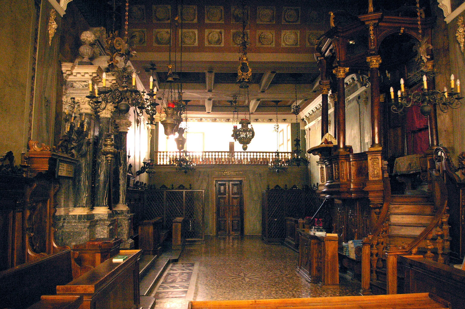Photograph of room with ornate columns and pews along both sides, a raised platform on the right side, and several chandeliers. 