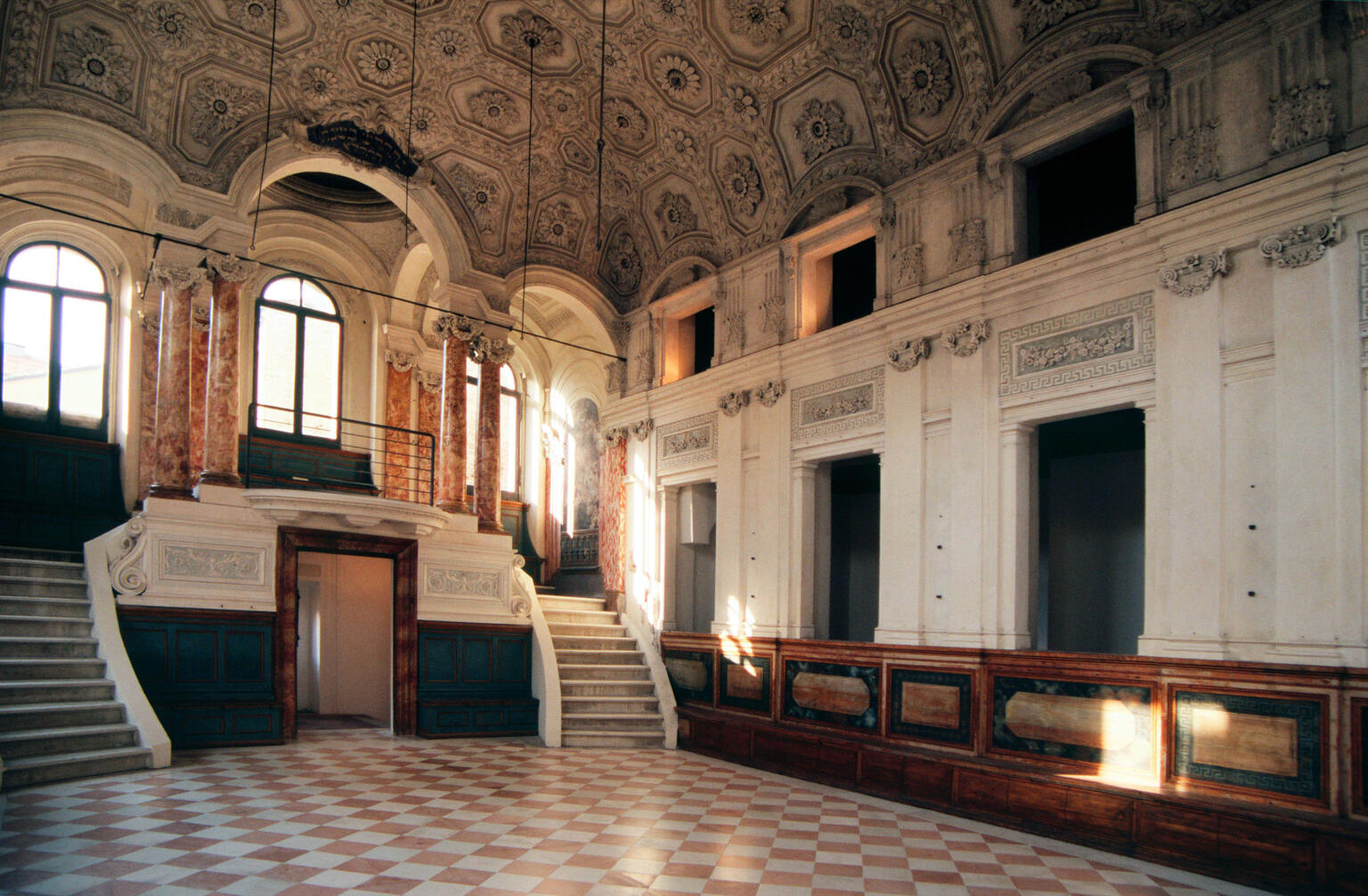 Interior of room with columns and short double staircase at one end and pews along wall.