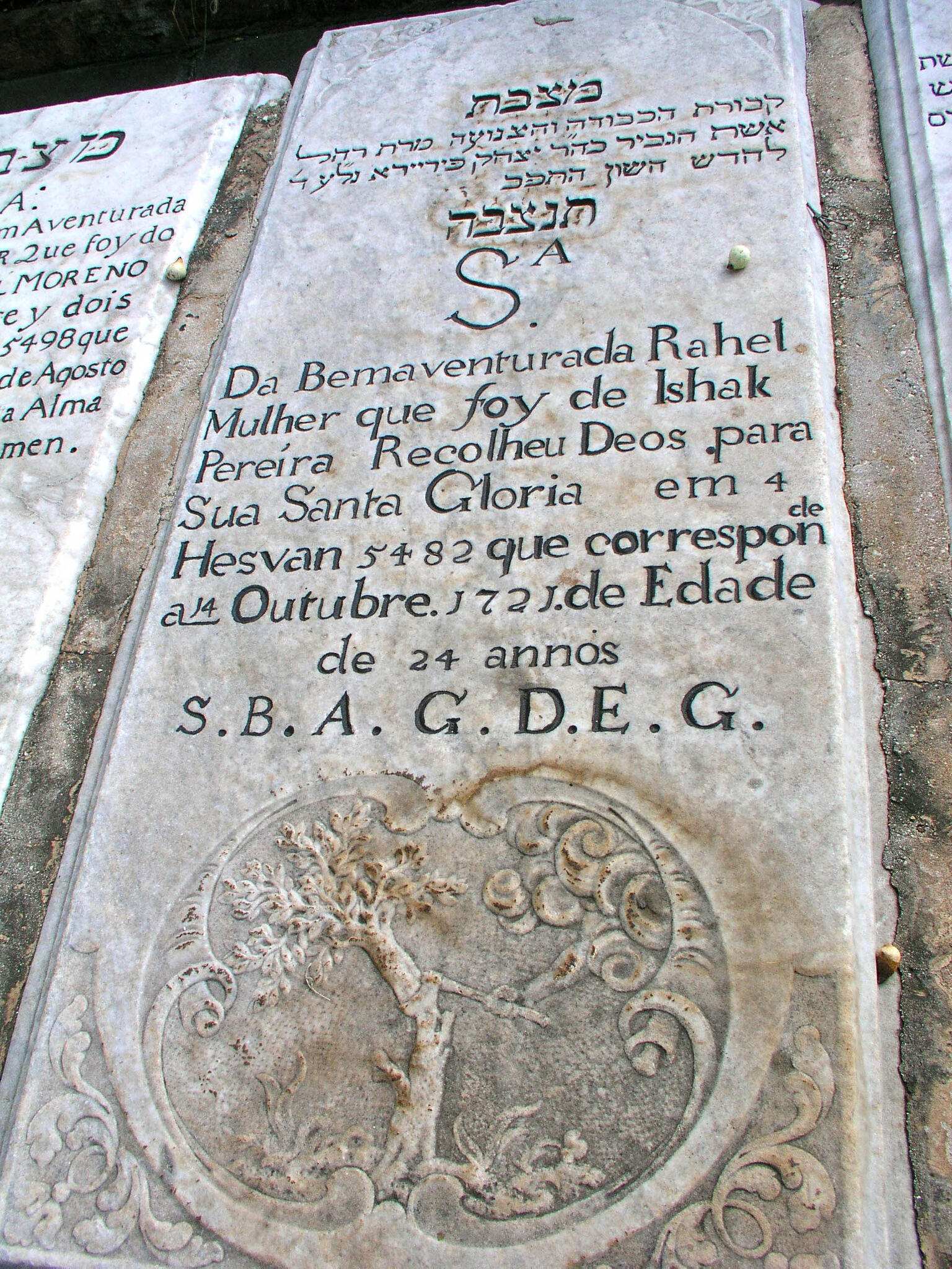 Tombstone with inscription in Hebrew and Portuguese above floral motifs and engraving of hand with an ax reaching out of cloud to chop down tree.
