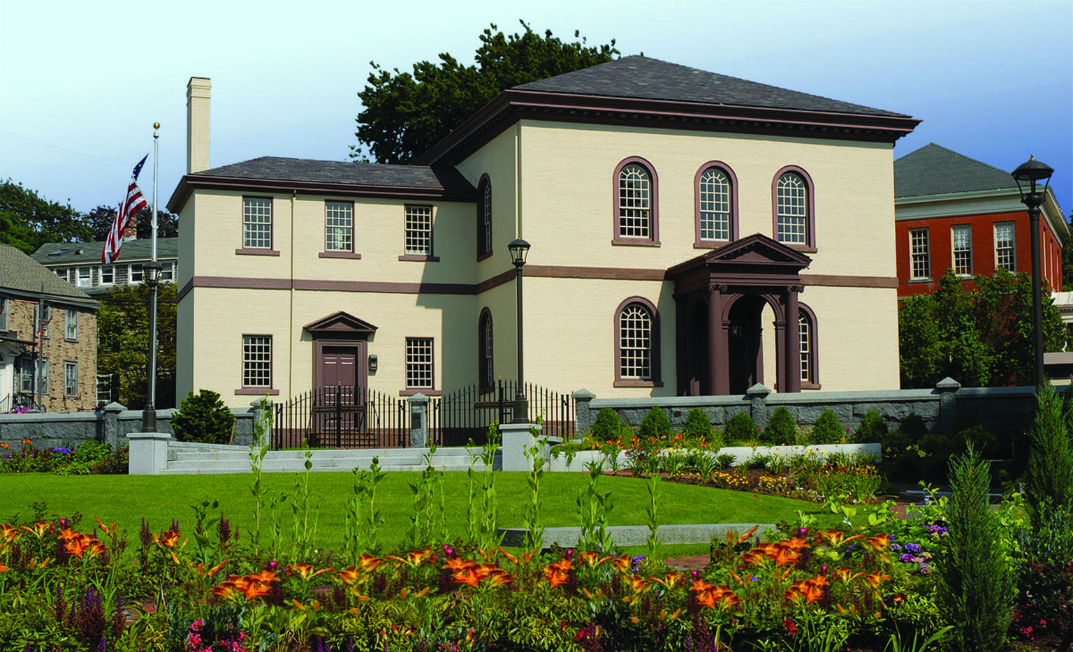 Photograph of exterior of building with semi-circular windows featuring American flag on left and flower garden in front.