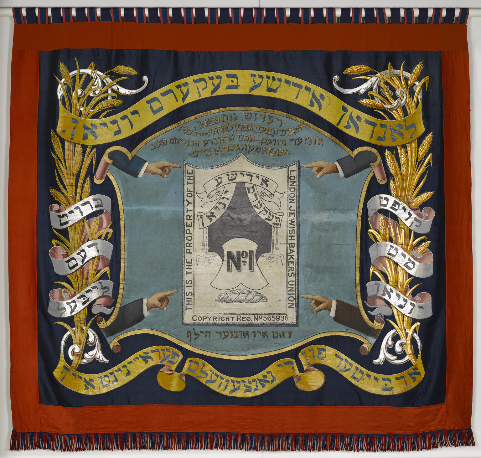 Banner with four arms pointed toward a crest in the center featuring a loaf of bread and English text, framed by Yiddish text and sheaves of wheat.