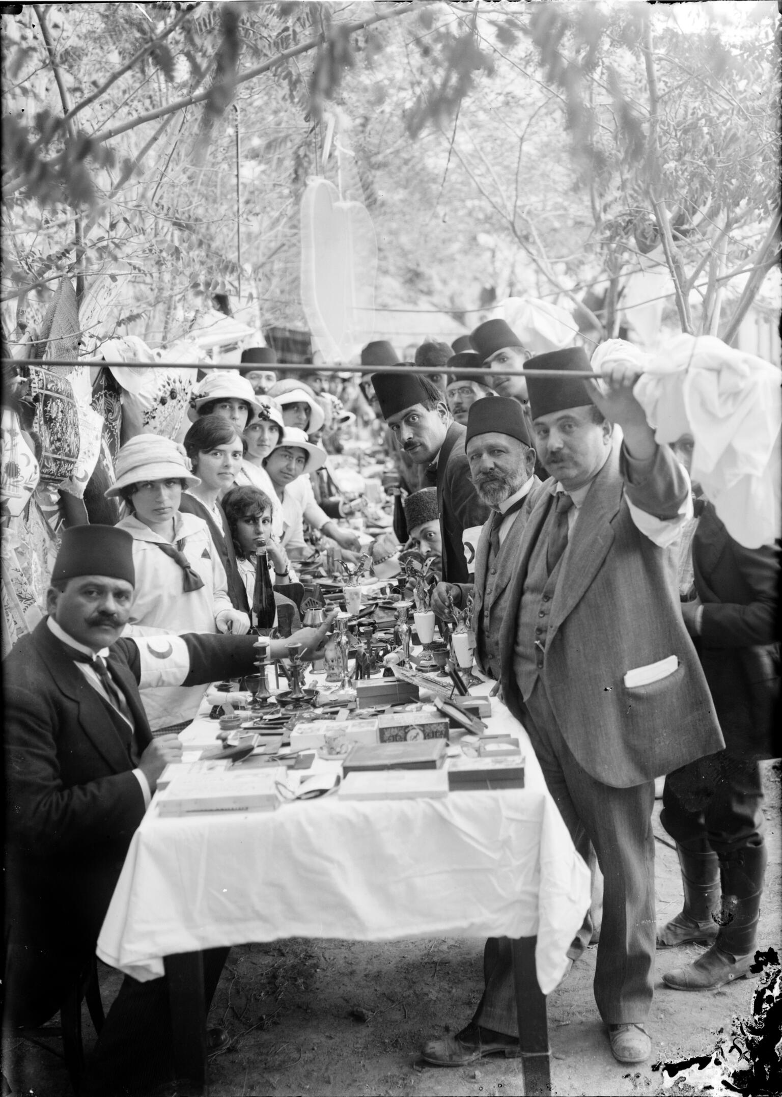 Photograph of women and men in fez hats sitting and standing at a long set table.