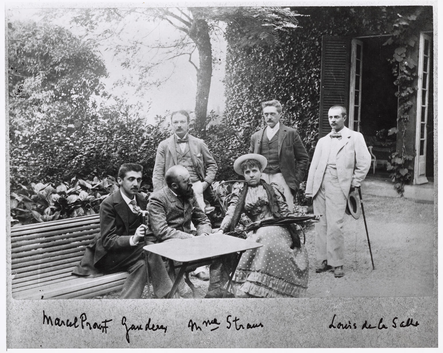 Photograph of several figures sitting at table in garden and signatures below as labels.