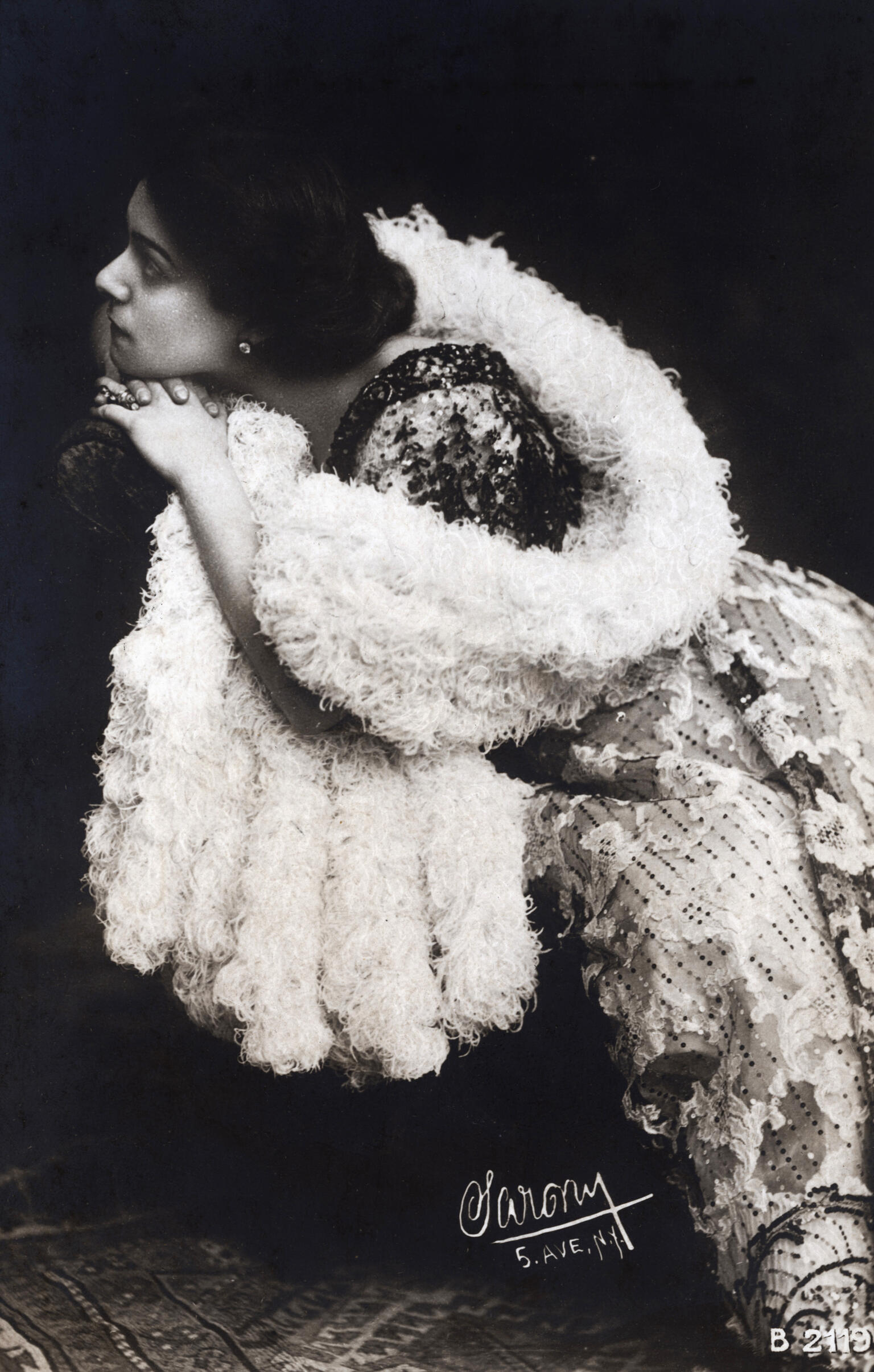 Photograph of woman in boa sitting on chair and looking to the side.