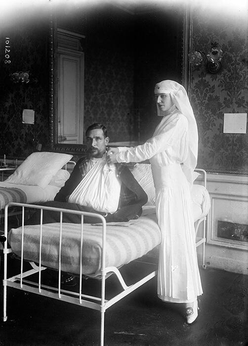 Photograph of woman in veil standing next to bed where man sits with arm in sling.