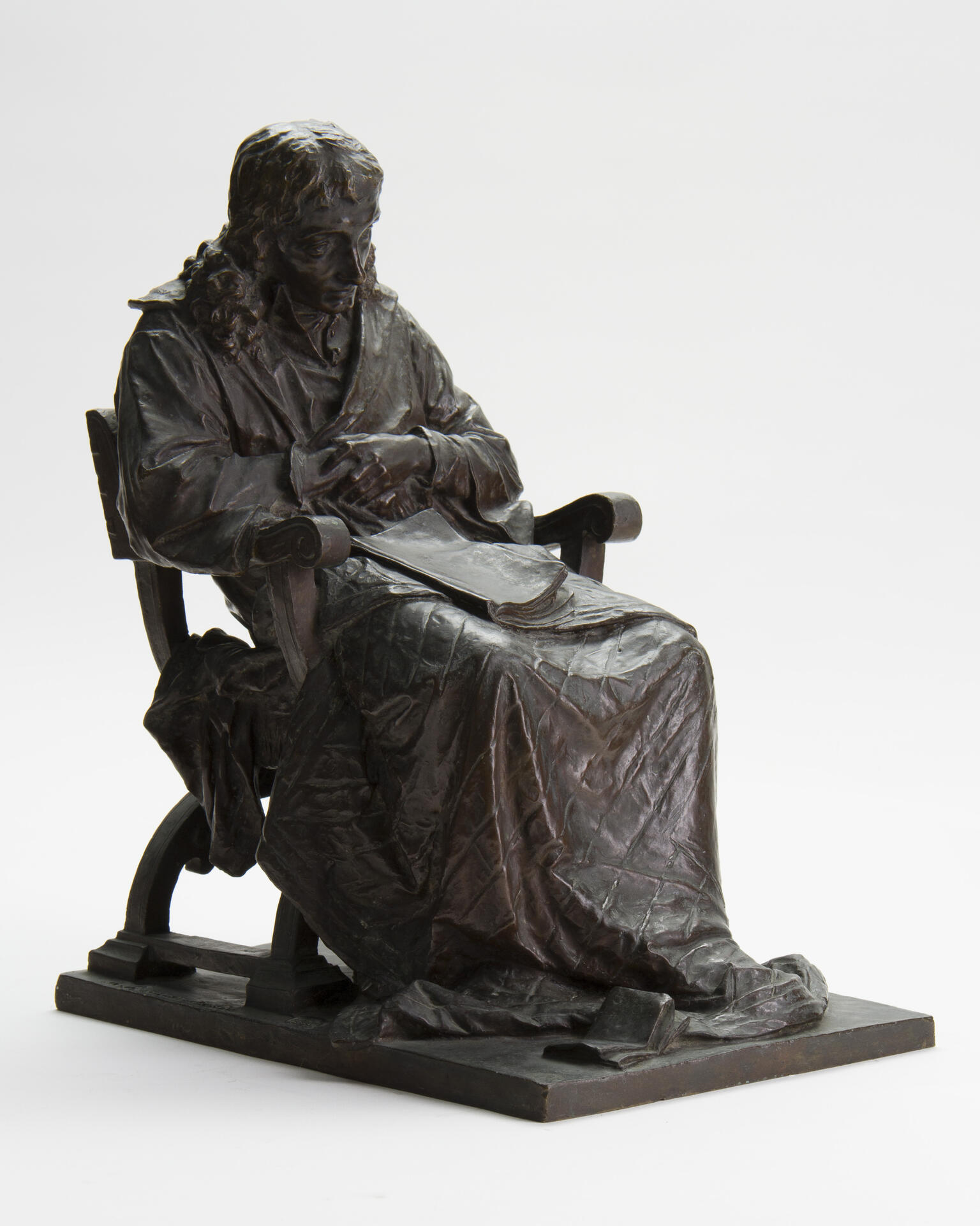Sculpture of seated figure with blanket draped over legs and book in lap.
