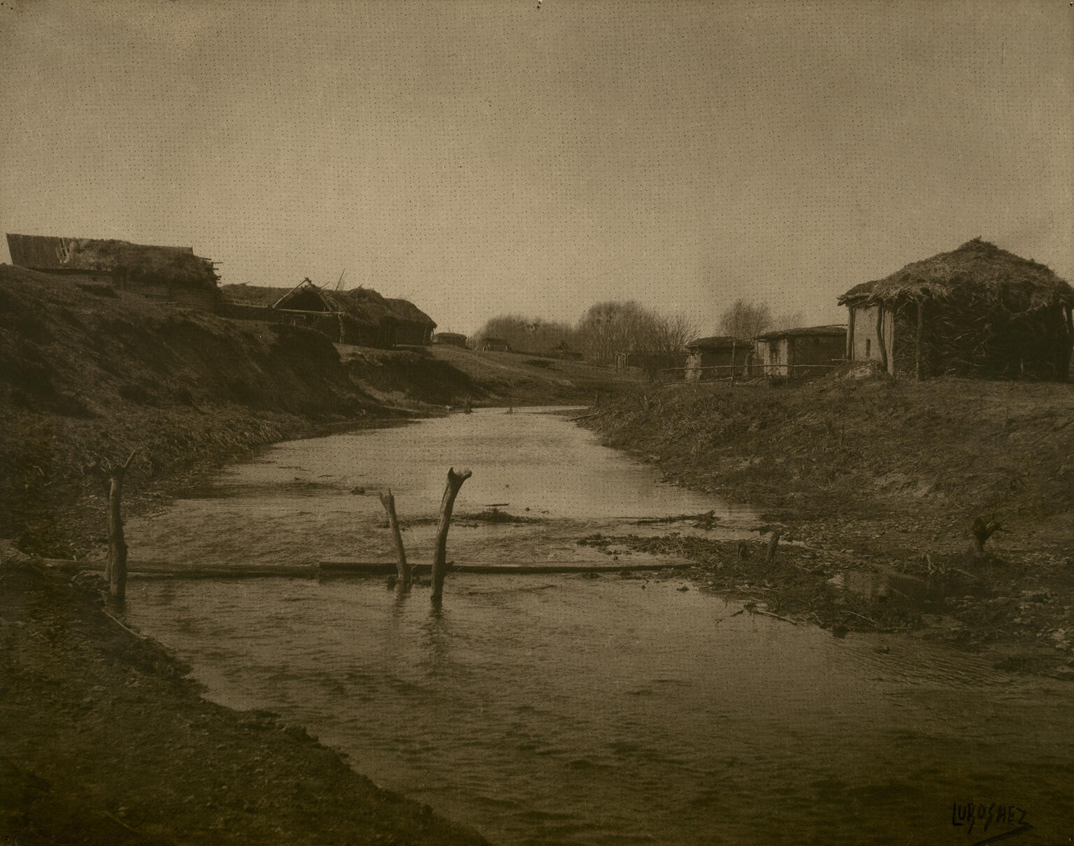 Photograph of landscape with creek lined with small huts. Photograph of landscape with creek lined with small huts.