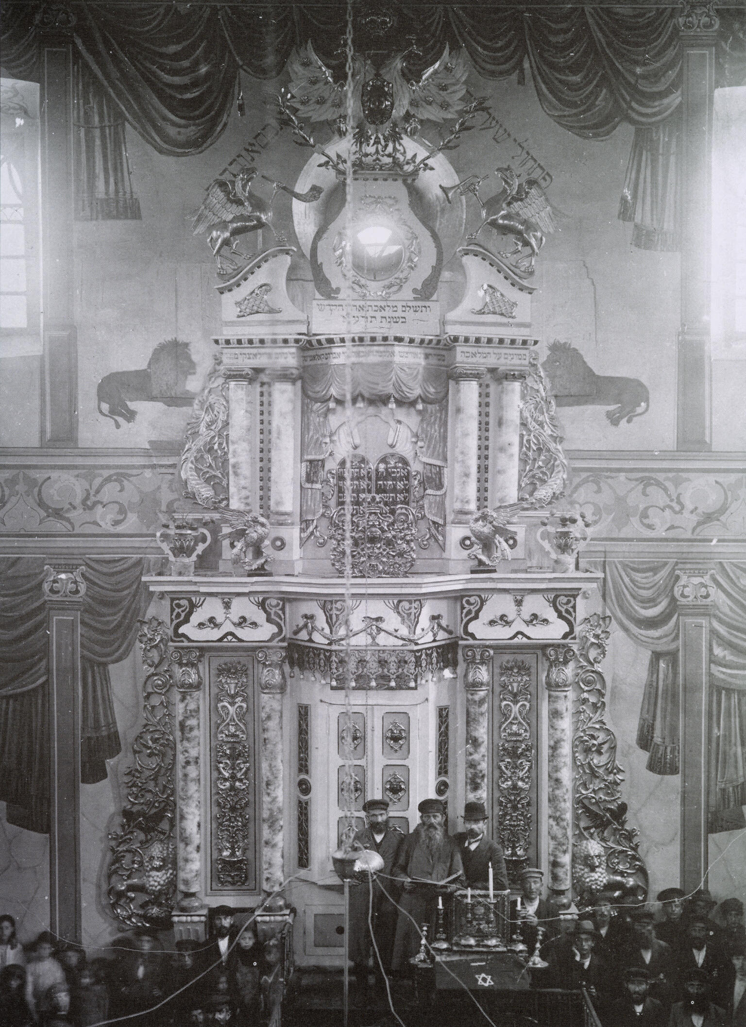 Photograph of large, ornate structure decorated with flourishes, lions, and vines, with three men in hats and beards standing in front of two closed doors at the bottom, and several other figures sitting in benches in foreground.