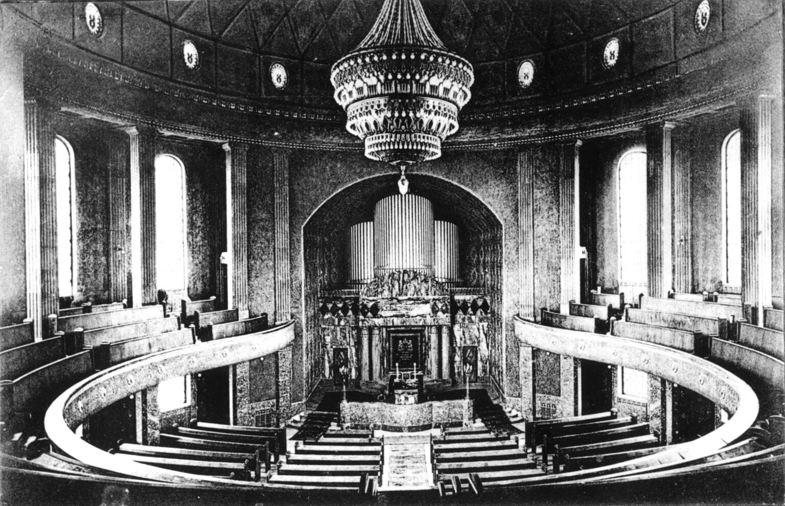 Photograph depicting organ and circular rows of benches, with large central chandelier.