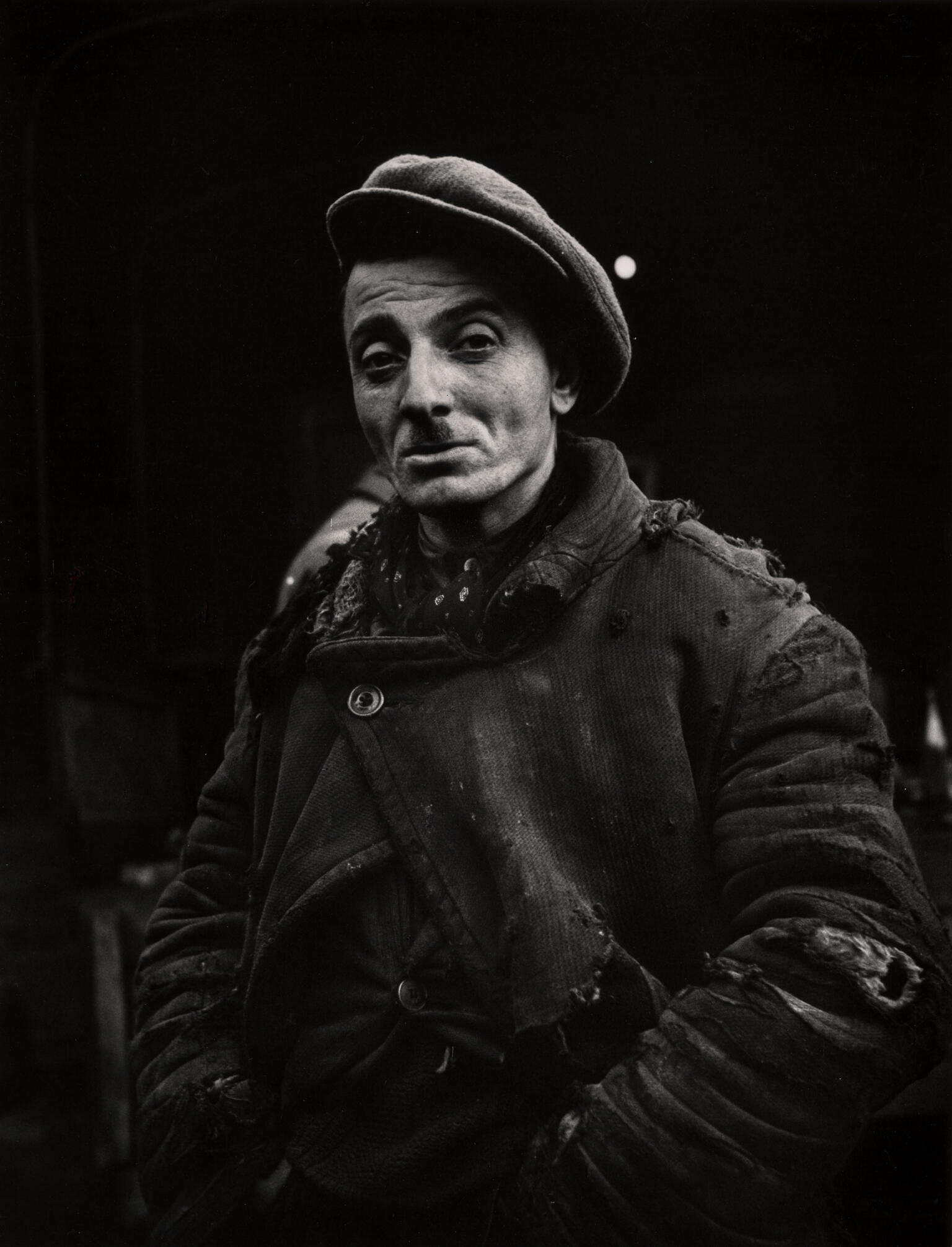 Photograph of man with mustache and hat wearing torn clothing with hands on hips, looking at camera. Photograph of man with mustache and hat wearing torn clothing with hands on hips, looking at camera.