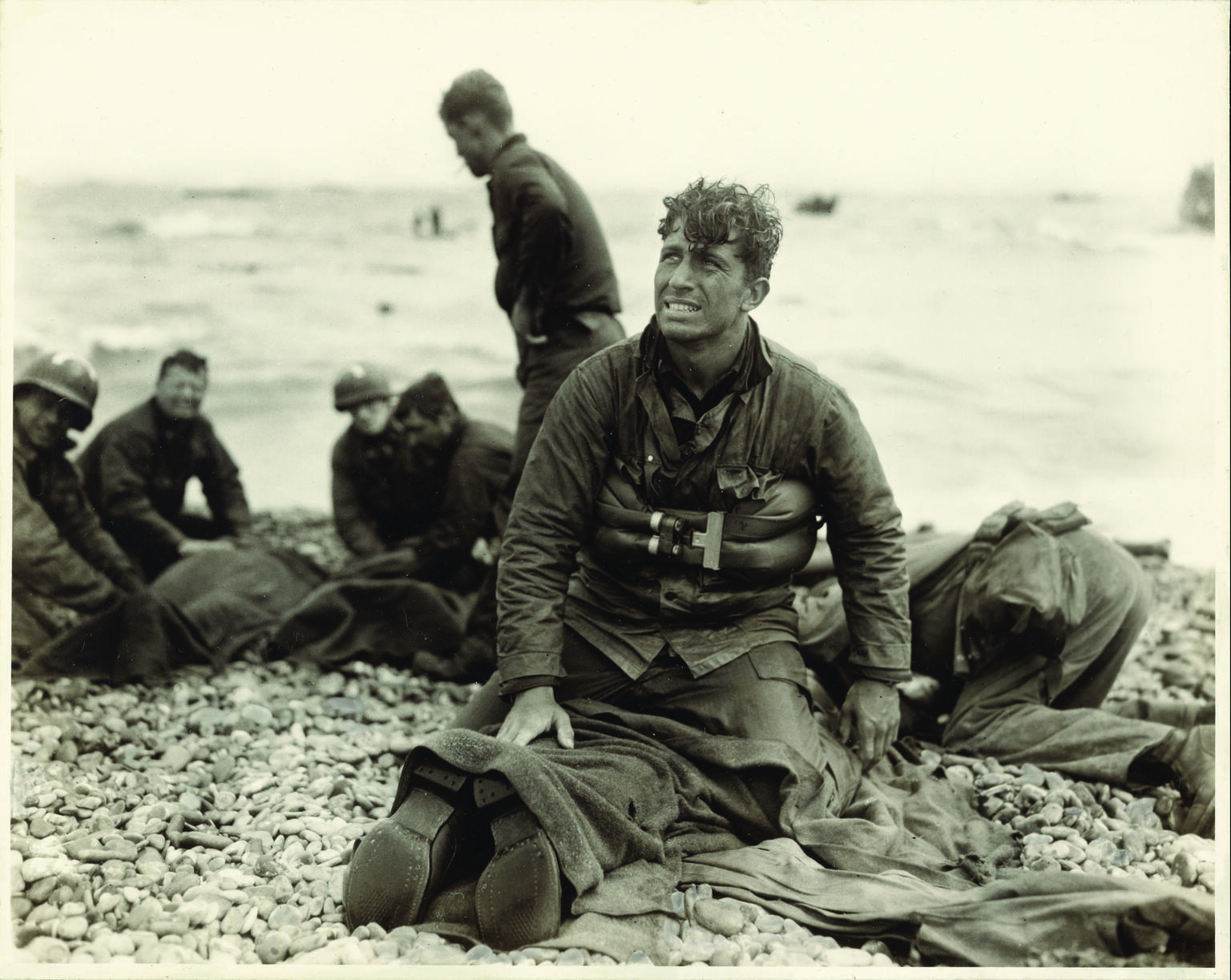 Photograph of soldiers on the beach, with man in center facing the viewer and looking up, as he kneels on top of a prone soldier. Photograph of soldiers on the beach, with man in center facing the viewer and looking up, as he kneels on top of a prone soldier.