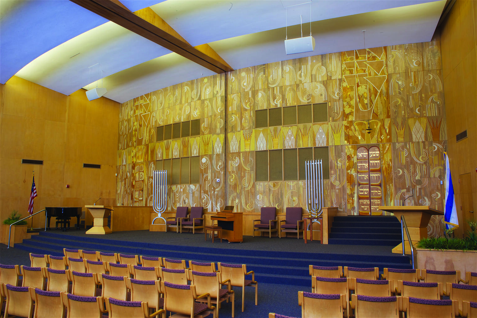 Photograph of interior view of a synagogue with rows of chairs facing a central platform featuring wall mural of painted panels with crowns, suns, Hebrew lettering, and spirals.
