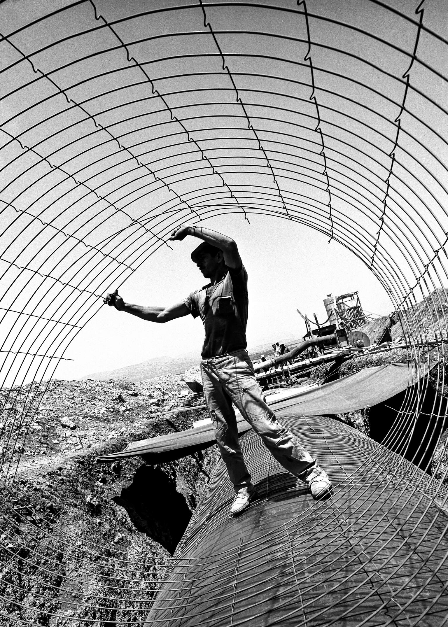 Photograph of man holding a wire cylinder and standing on a large pipe, with a construction site behind him. Photograph of man holding a wire cylinder and standing on a large pipe, with a construction site behind him.