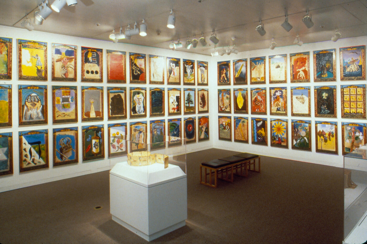 Installation photograph featuring walls of paintings and jewel case and bench in center of room.