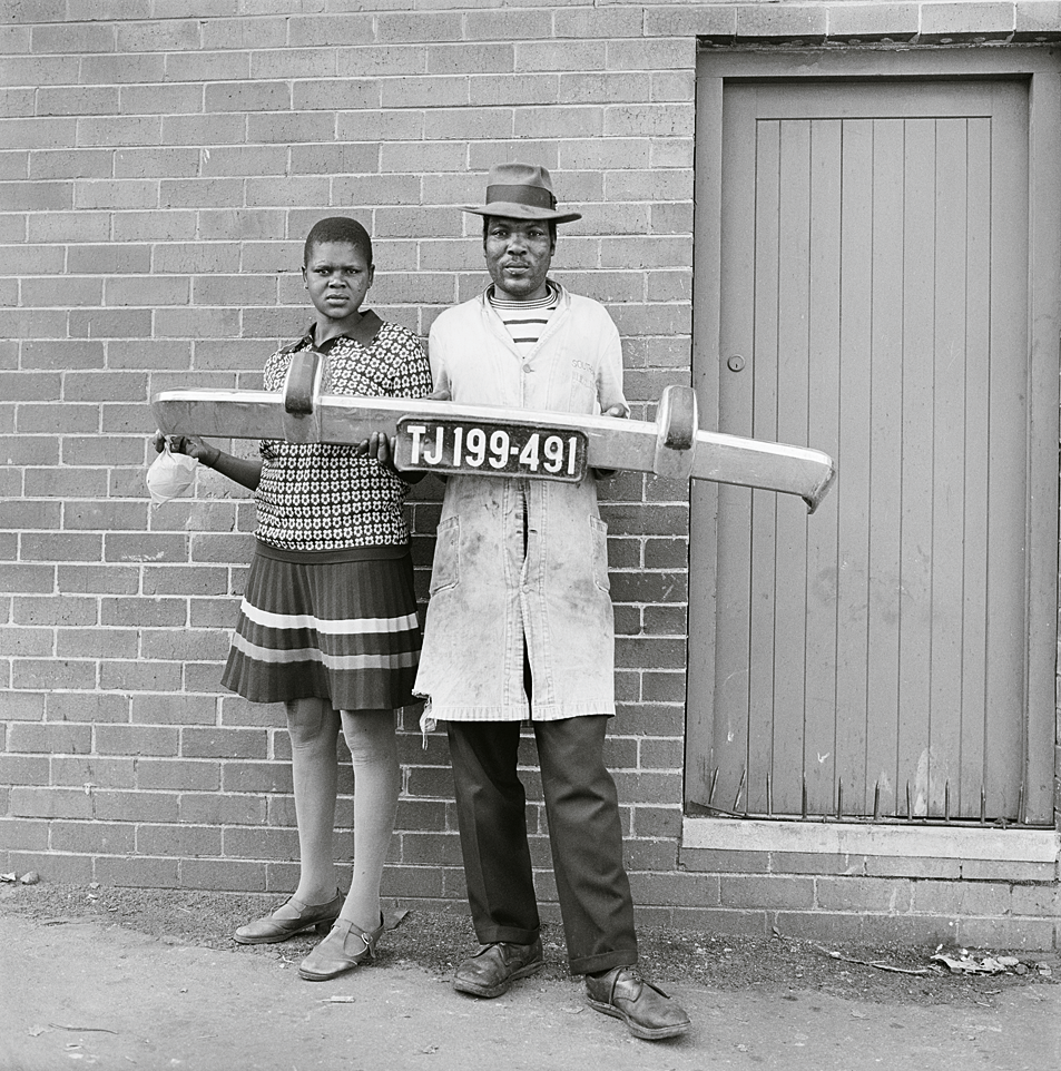 Photograph of man and woman standing and holding a car bumper in front of a brick wall.  