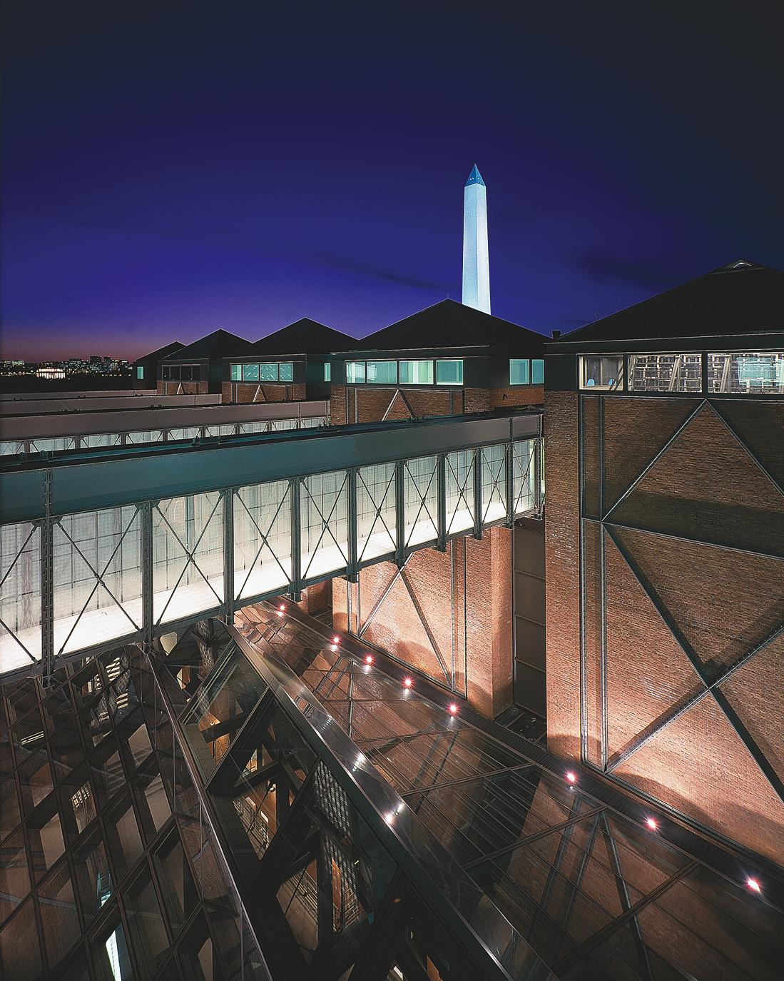 Photograph of brick building exterior with skywalks over a smaller building below with a metal and glass roof and a tall monolith in the background. Photograph of brick building exterior with skywalks over a smaller building below with a metal and glass roof and a tall monolith in the background.