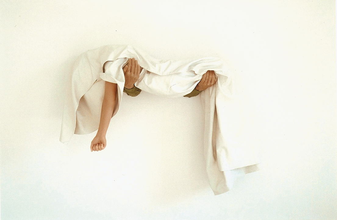 Art installation of arms coming out of a wall holding body covered in sheet with one arm exposed. 