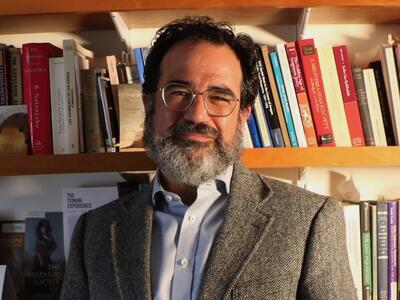 A man with short dark hair, graying beard, glasses, a light blue shirt, and a tweed blazer looks at the camera; a shelf of books is behind him.