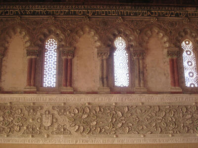 Photo of a wall with a row of ornate arches and windows and a row of ornate floral decorative engraving, each surrounded by three lines of engraved Hebrew text.