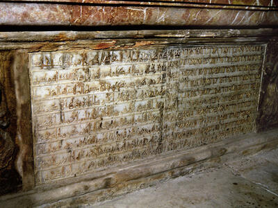 Photo of white stone relief in the bottom of a wall of gold lettering in multiple languages, with a column of repeated castles and lions down the middle.