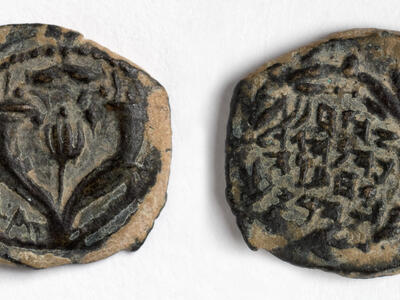 Two sides of an ancient, worn coin. One side depicts a pomegranate flanked by horns; the other side features several lines of inscription in Paleo-Hebrew script.